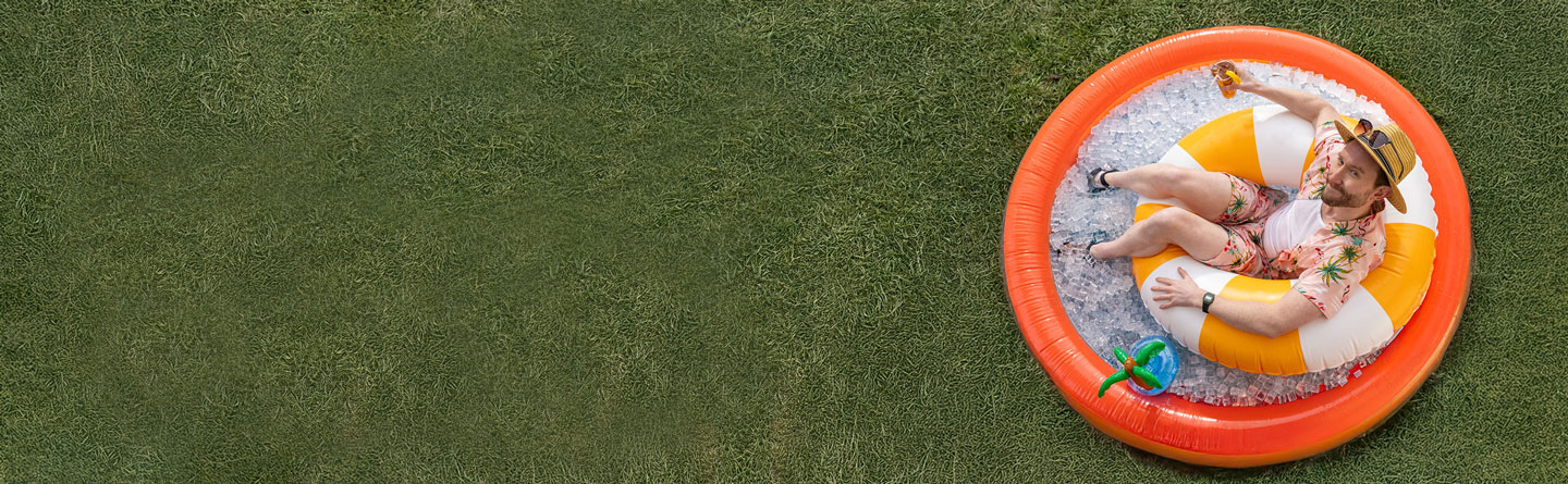Person holding a beverage wearing summer clothes inside of a small inflatable pool full of ice outside on green grass