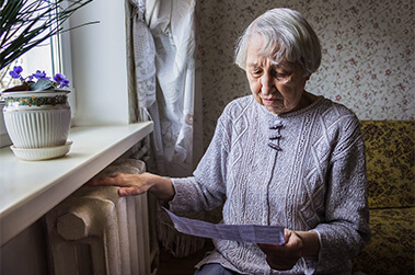 Elderly lady looking at statement next to heater in her home, trying to stay warm.